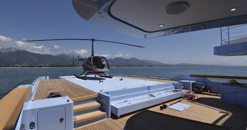 A small helicopter is parked on the deck of a yacht at sea, with mountains and coastline visible in the background.